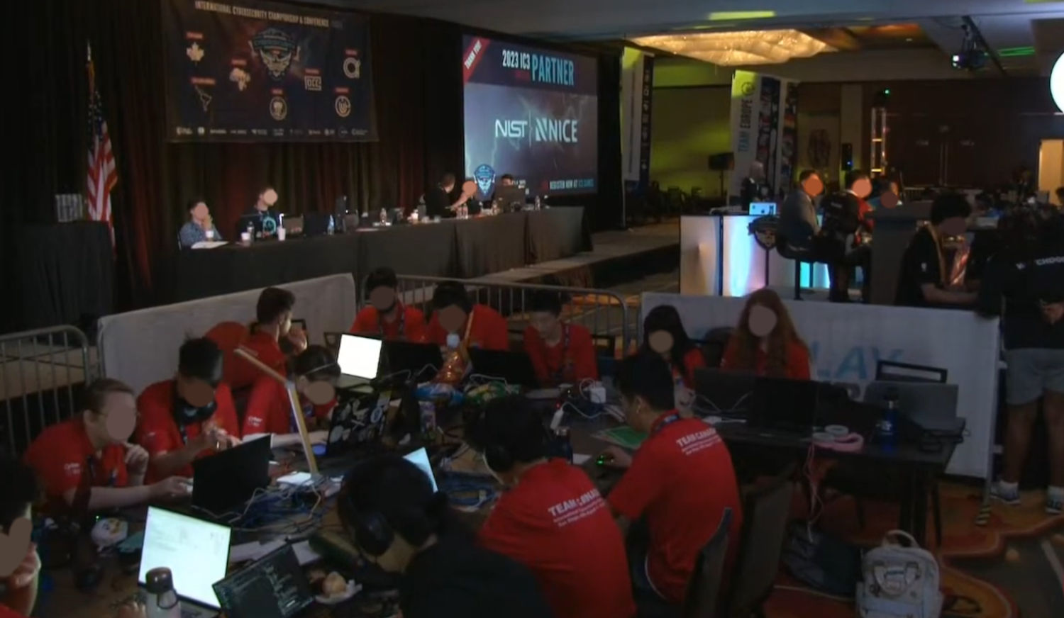 Low resolution photo of the competition 'arena', showing team Canada and the announcers. The room is large and dim, lit primarily by moving spotlights.